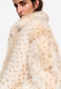Abrigo de piel sintética color beige claro con una textura suave y esponjosa y un patrón de manchas más oscuras, con un cuello alto y un corte holgado.