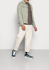 Ljusgrön denimjacka, brun t-shirt, krämfärgade byxor och gröna sneakers med vita accenter. Slim fit, uppvikta ärmar.