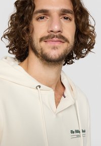 Kremkrāsas kapučjaka, izgatavota no mīksta auduma, ar priekšējo kabatu, auklu kapuci un zaļiem uzrakstiem uz krūtīm, papildināta ar akcentiem.