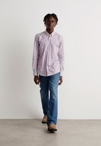 Camisa de manga larga a rayas en púrpura claro y blanco, combinada con jeans azules y zapatos beige. Modelo caminando sobre un fondo neutral.