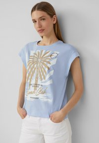 Błękitny bawełnowy t-shirt z krótkim rękawem, z grafiką palmy i napisem, o luźnym kroju. Stylizowany z białymi spodniami.