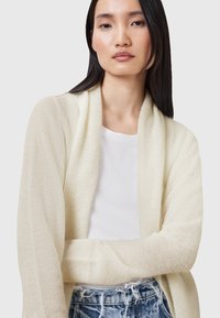 Svetlá béžová kardigán s textúrovaným pleteným dizajnom, otvorený predný strih, dlhé rukávy a voľný strih, nosený cez biely top a roztrhnuté džínsy.