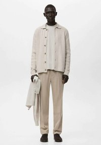 Beżowy sweter z dzianiny na jasnoszarej koszuli, w połączeniu z beżowymi spodniami. Model trzyma pasujący sweter, podkreślając neutralną paletę kolorów.