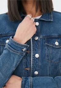 Veste en denim bleu foncé avec des boutons en argent, des poches poitrine et des détails de couture visibles. Le tissu semble légèrement usé avec une texture lisse.