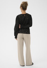 Göndör hajú nő, aki fülbevalót visel, fekete csipkés, hosszú ujjú felsőt és bézs, széles szárú nadrágot hord, és kezét a háta mögött tartja.