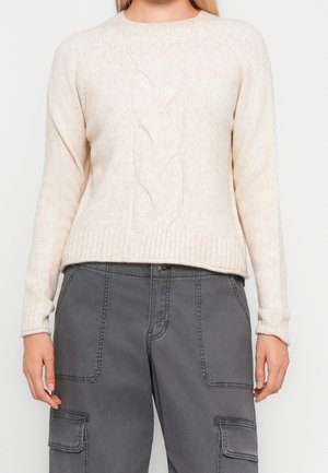 Kvinde iført en cremefarvet kabelstrikket sweater kombineret med højtaljede grå cargobukser med forlommer og sidelommer.