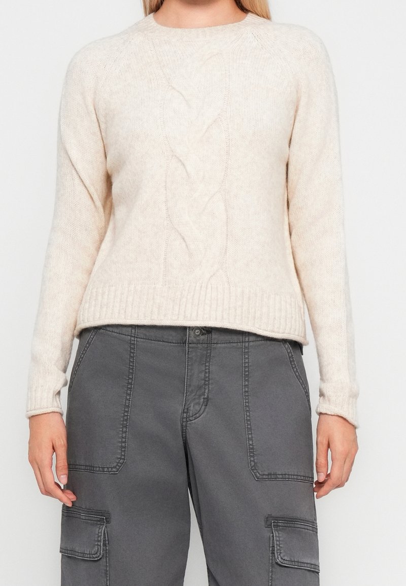 Femme portant un pull en maille torsadée crème associé à un pantalon cargo gris taille haute avec poches avant et rabats latéraux.