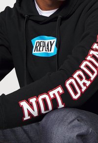Persona vestida con una sudadera negra con el logo "REPLAY" en el pecho y el texto "NOT ORDINARY" en blanco con contorno rojo a lo largo de la manga.
