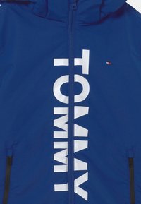 Blauwe zip-up jack met grote witte "TOMMY" tekst. Heeft een capuchon, twee zijzakken met rits en een klein rood logo. Glad textuur.