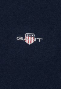 Tessuto blu navy con una texture morbida, caratterizzato da un logo: uno scudo con strisce rosse e bianche e tre stelle bianche sopra la parola "GANT."