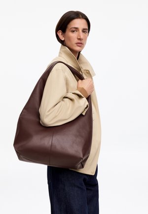 Persona con giacca beige tiene una grande borsa a tracolla in pelle marrone, in piedi davanti a uno sfondo bianco uniforme.