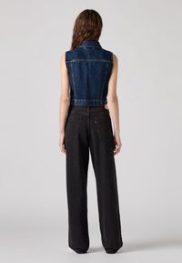 Personne aux cheveux longs portant un gilet en denim sombre sans manches et un jean noir ample, debout, dos tourné, sur un fond gris uni.