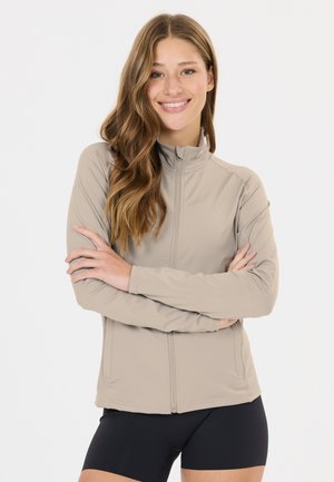 Junge Frau mit langen, welligen Haaren, lächelnd, trägt eine beige Jacke mit Reißverschluss und schwarze Shorts, steht mit verschränkten Armen vor einem weißen Hintergrund.