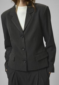 Ženska, oblečena v temno prugasto blazer s tremi gumbi in izrezom, čez belo pleteno pulover, z rokami v žepih ustreznih hlač.