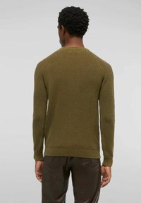 Uomo con capelli ricci corti, che indossa un maglione a maglia verde oliva e pantaloni marroni scuri, in piedi con la schiena rivolta verso una sfondo neutro.