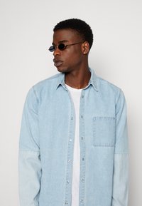 Camicia di denim azzurro chiaro con maniche lunghe, caratterizzata da una chiusura con bottoni, tasca sul petto e colore e texture delle maniche in contrasto. Occhiali da sole ovali neri.
