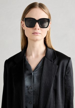 Ochelari de soare negri, supradimensionați, cu o ramă masivă și lentile întunecate, asortați cu o cămașă gri satinat cu nasturi și un sacoș negru.