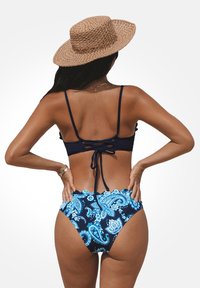 Navyblaues Bikini-Set mit einem gebundenen Oberteil und hochgeschnittenem Unterteil mit einem blauen Paisleymuster. Ein geflochtener Strohhut ergänzt das Design.
