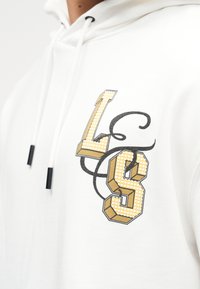 Sweat à capuche blanc avec cordons, présentant de grandes lettres stylisées en or et blanc "L" et "S" reliées par des arabesques décoratives noires sur la poitrine.
