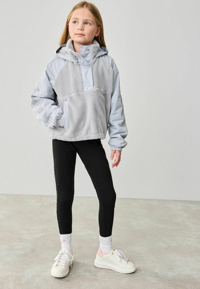 Grå fleece och vindjacka, stor framficka, med svarta leggings och vita sneakers med rosa detaljer, står på ett ljust golv.