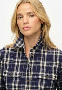 Navy and cream plaid flannel shirt with a collar, two front pockets, and button closures. Material appears soft with a textured finish.