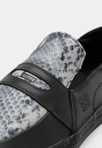 Κοντινό πλάνο μαύρου παπουτσιού χωρίς κορδόνια με γκρι επάνω μέρος σε σχέδιο φίδι και λογότυπο "Globe" στη λωρίδα που διασχίζει την κορυφή.