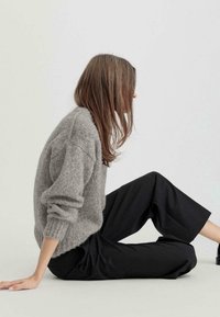 Suéter de punto gris con puños acanalados, combinado con pantalones de pierna ancha negros. Postura sentada, textura suave y corte relajado. Fondo neutro.