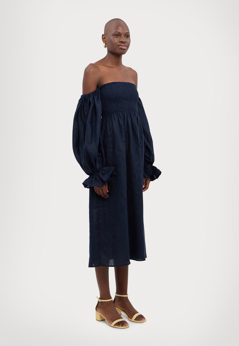 Marineblå off-shoulder kjole med smocket liv og voluminøse lange ermer. Har volanger i mansjettene og ankel-length snitt, parret med gule blokkhæl sandaler.