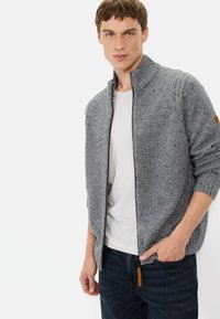 Grijze gebreide cardigan met een hoge kraag, ritsen en een textuurpatroon. Kenmerkt een leren accent en geribbelde manchetten. Gecombineerd met een wit T-shirt.