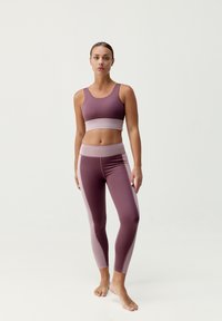 Sportovní podprsenka a legíny v tmavě mauve a světle fialové barvě. Materiál vypadá hladce s kontrastními bočními panely a širokým páskem.