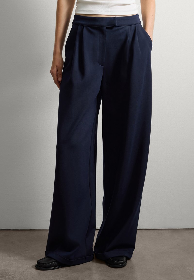 Personne portant un pantalon large bleu marine, les mains dans les poches et des chaussures noires, se tenant devant un mur clair.