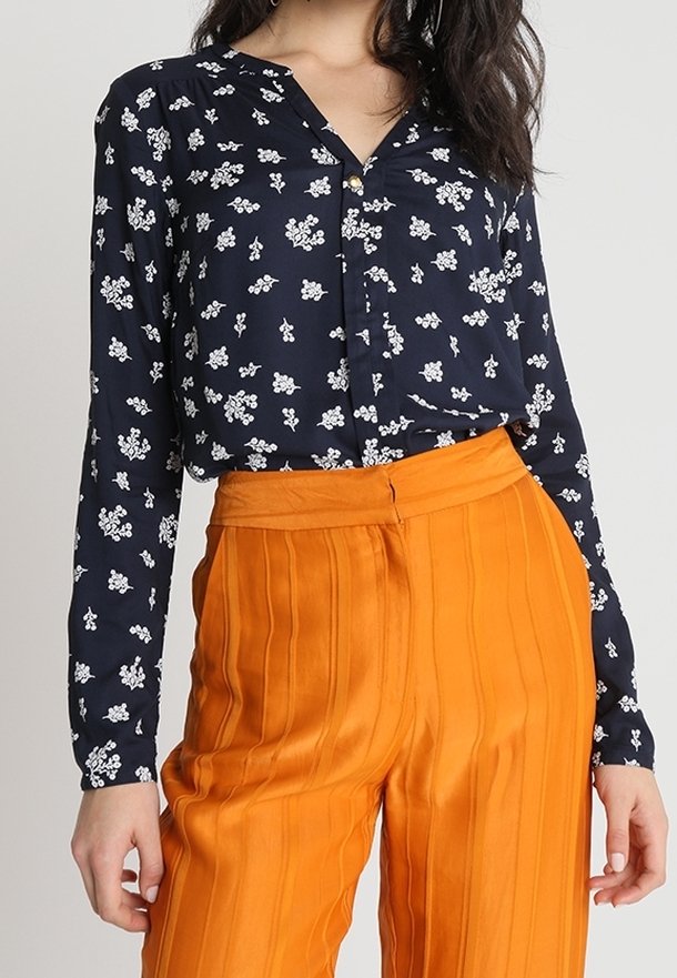Femme portant un chemisier bleu marine à manches longues avec des imprimés floraux blancs et un pantalon plissé taille haute de couleur orange vif.