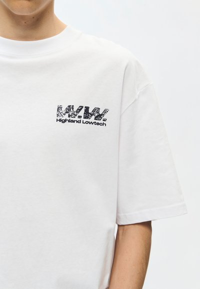 Άτομο που φοράει λευκό T-shirt με μαύρο κείμενο "W.W. Highland Lowtech" τυπωμένο στην πάνω αριστερή περιοχή του στήθους.