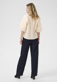 Blouse en beige clair texturé avec des manches courtes bouffantes, associée à un pantalon large bleu foncé et des bottes à talons noires. Design simple à l'arrière.