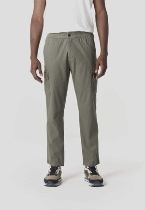 Olívazöld cargo nadrágot és bézs, fekete-fehér sportcipőt viselő férfi áll egy egyszínű világosszürke háttér előtt.