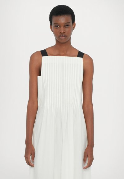 Weißes ärmelloses Kleid mit vertikalen Falten und schwarzen Schulterträgern. Der Stoff ist leicht mit einer glatten Textur und einem fließenden Design.