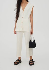 Conjunto de dos piezas de canalé en color crema que incluye una blusa sin mangas con botones de madera y pantalones cortos. Combinado con un pequeño bolso negro y sandalias planas.