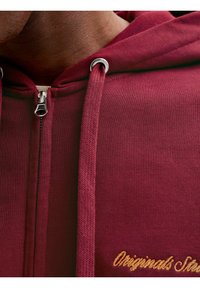 Hoodie bordeaux zippé avec fermeture éclair argentée, tissu texturé, cordons de serrage, et "Originals" brodé en or sur le devant.