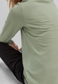 Top a maniche lunghe in morbido tessuto verde chiaro, con un fit rilassato, dotato di un collo rotondo e spacchi laterali. Indossato con pantaloni neri.