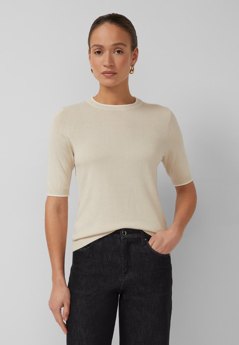 Žena u bež džemperu s kratkim rukavima i crnim trapericama, stoji s jednom rukom u džepu, ispred jednobojne sive pozadine.