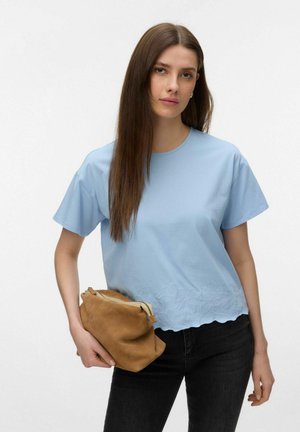 Ung kvinde med langt brunt hår iført en lyseblå broderet kortærmet bluse og sorte bukser, der holder en lille brun ruskinds taske.