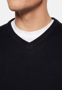 Czarny sweter z dekoltem w serek z prążkowanym wykończeniem, noszony na białej koszulce z okrągłym dekoltem, wykonany z gładkiego, teksturowanego dzianinowego materiału.