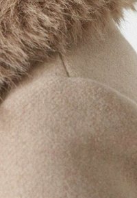 Beige villakangastakki, jossa on pehmeä, teksturoitu tekoturkiskaulus. Kankaan pinta on sileä ja hiukan karhea, korostaen luonnollisia värimuutoksia.
