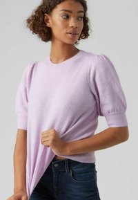 Lilla genser med korte pufferm, ribbestrikket hals og kant, laget av myk, teksturert stoff. Kombinert med mørke denimjeans.