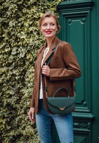 Grüne Lederhandtasche mit goldenen Akzenten, strukturierter Form und einem oberen Griff. Das Modell trägt einen braunen Wildlederblazer und helle Jeans.