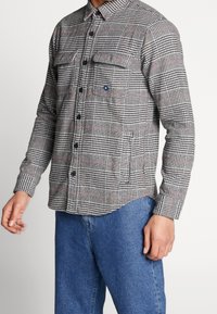 Man som bär en rutig skjorta i svart, vitt och rött med knappknäppning och fickor med lock, som är kombinerad med blå jeans, stående mot en enkel vit bakgrund.