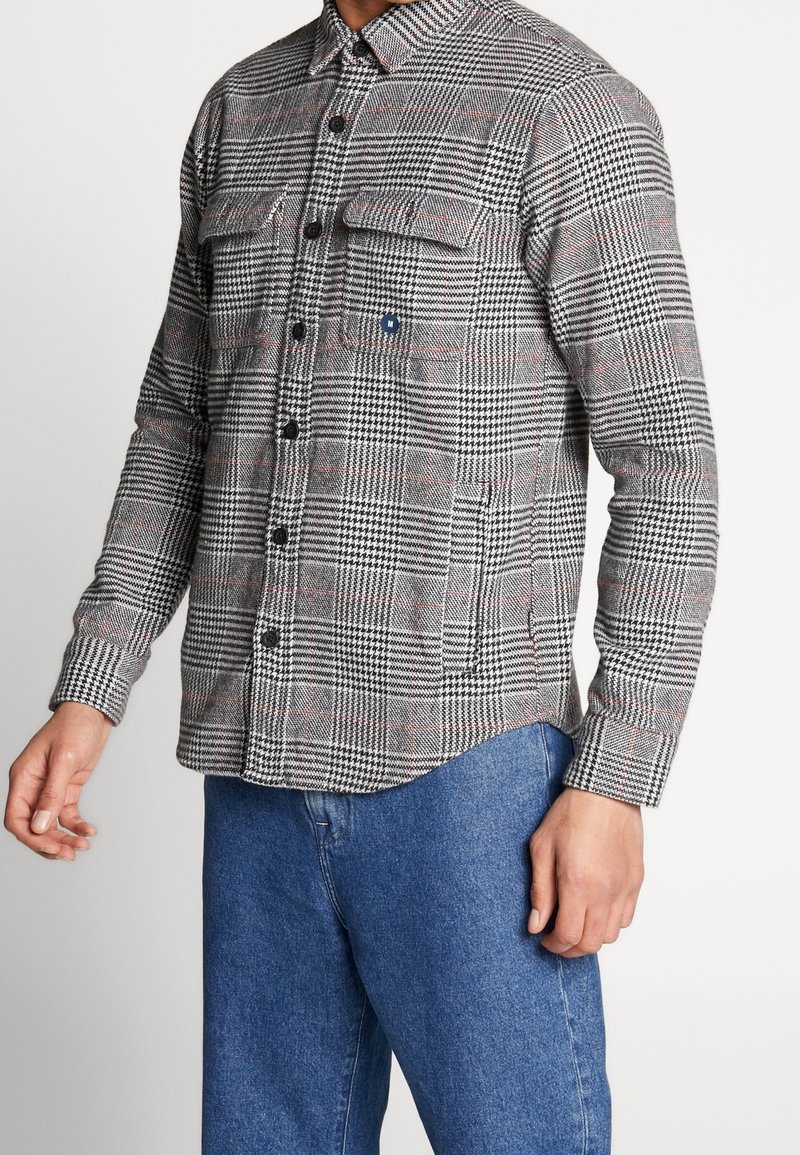 Man som bär en rutig skjorta i svart, vitt och rött med knappknäppning och fickor med lock, som är kombinerad med blå jeans, stående mot en enkel vit bakgrund.