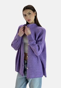 Cardigan lavorato a maglia viola con colletto alto e bottoni frontali, caratterizzato da una vestibilità ampia e tessuto texturizzato. Indossato sopra una maglietta bianca.