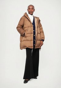 Osoba oblečená v béžovom zateplenom kabáte, námorníckom svetri s gombíkmi, bielom roláku, čiernych nohaviciach s širokými nohavicami a čiernych topánkach na bielom pozadí.