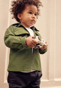 Jeune enfant aux cheveux bouclés portant une veste verte et un pantalon noir, tenant un appareil photo jouet, regardant pensivement sur le côté.
